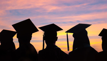 New graduates wearing academic caps are visible in silhouette against a colorful sunrise.